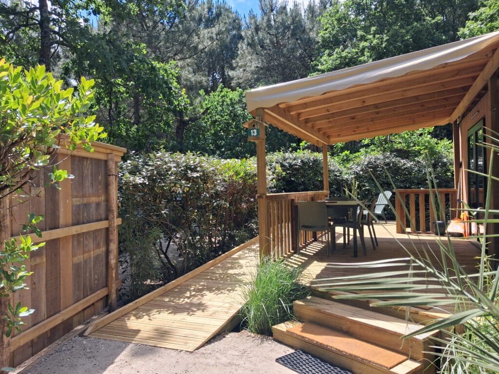 Terrasse en bois ombragée n°13 avec rampe d'accès, table de jardin et végétation environnante.