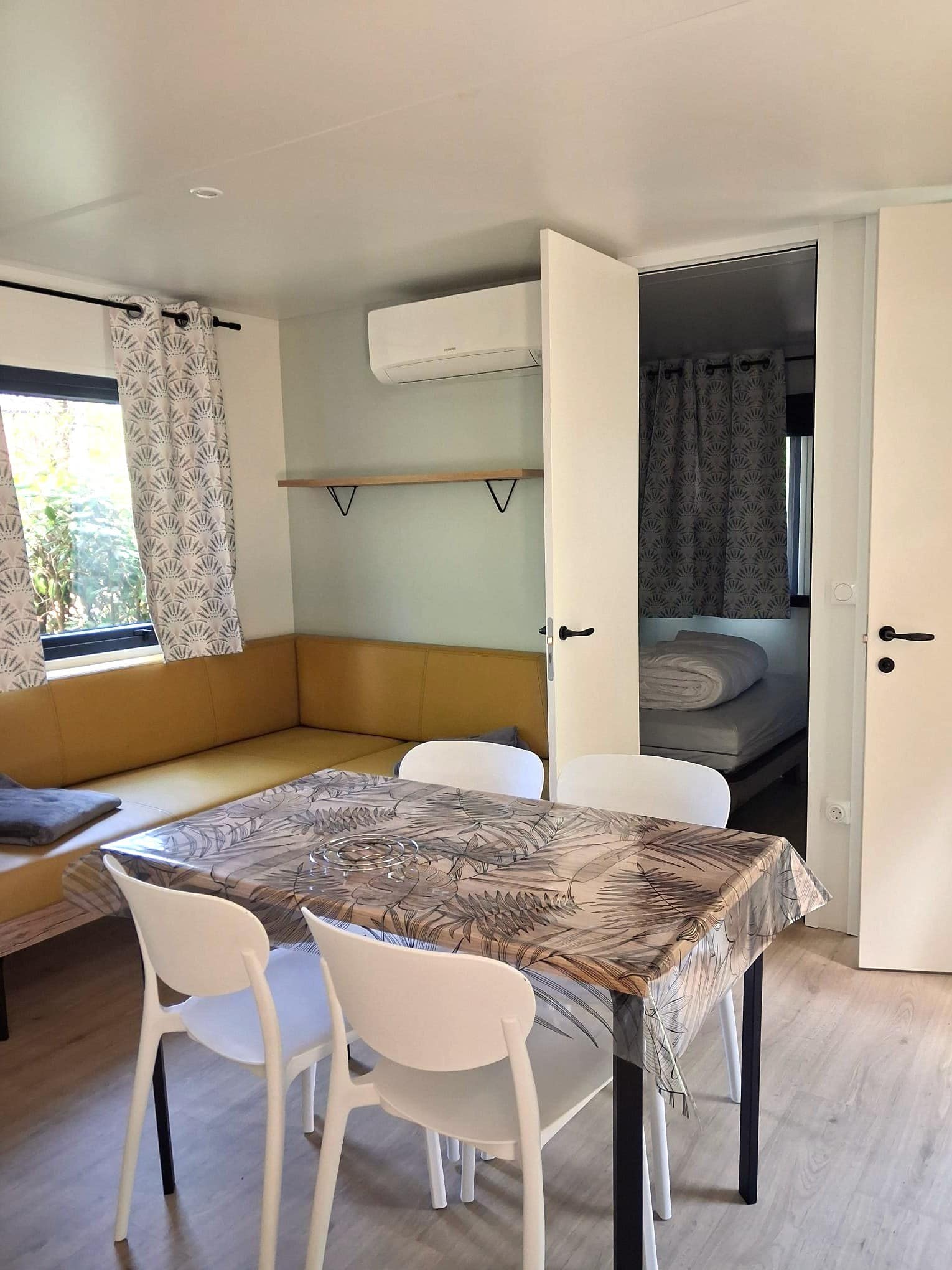 Salon de mobil-home moderne avec table à manger, banquette jaune, chaises blanches et aperçu d'une chambre lumineuse.
