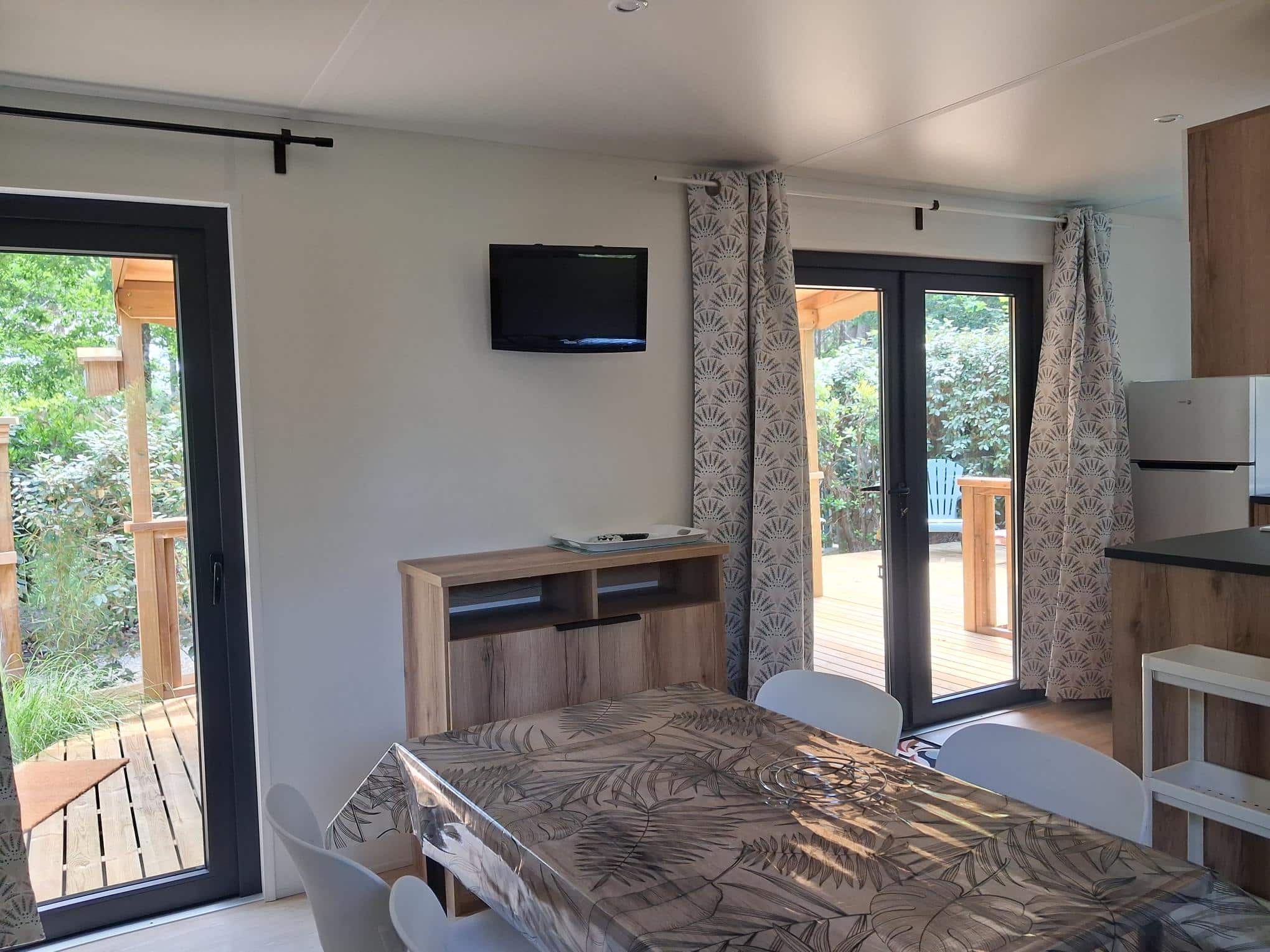 Intérieur de mobil-home moderne avec coin repas, salon TV et accès direct vers une terrasse en bois ensoleillée.