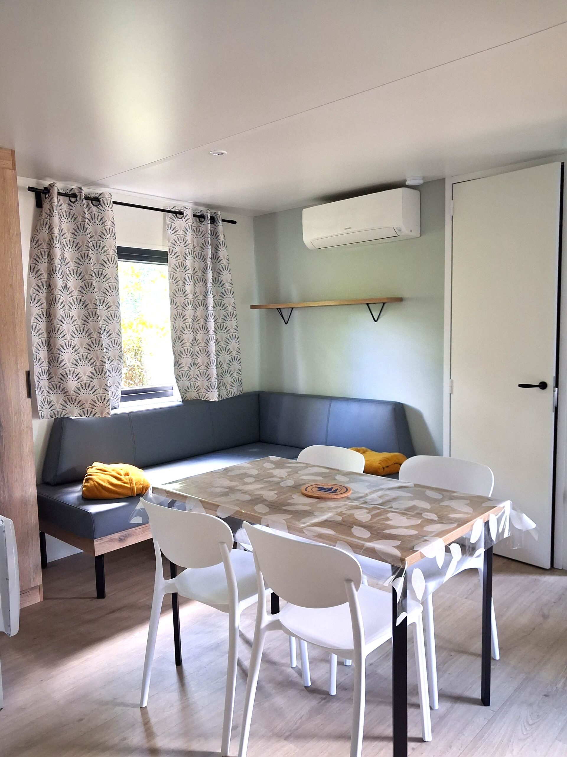 Coin repas moderne d'un mobil-home avec banquette grise, table en bois, chaises blanches et climatisation.