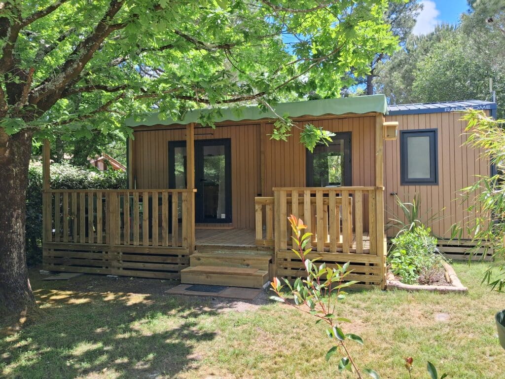 Mobil-home Rapid Home en bois avec terrasse ombragée et jardinet dans un camping verdoyant.