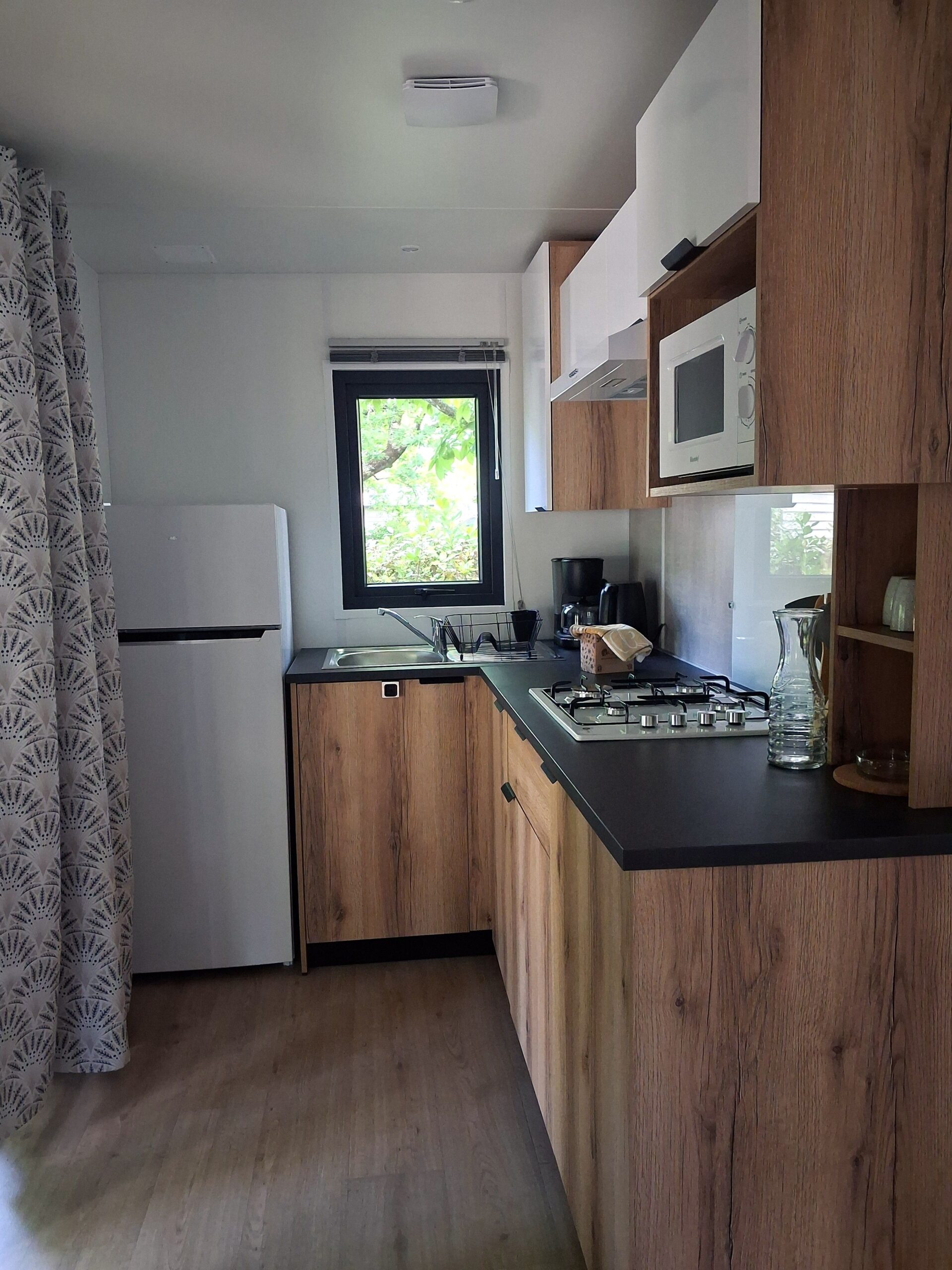 Petite cuisine moderne avec meubles en bois, plan de travail noir, évier sous fenêtre et réfrigérateur blanc.