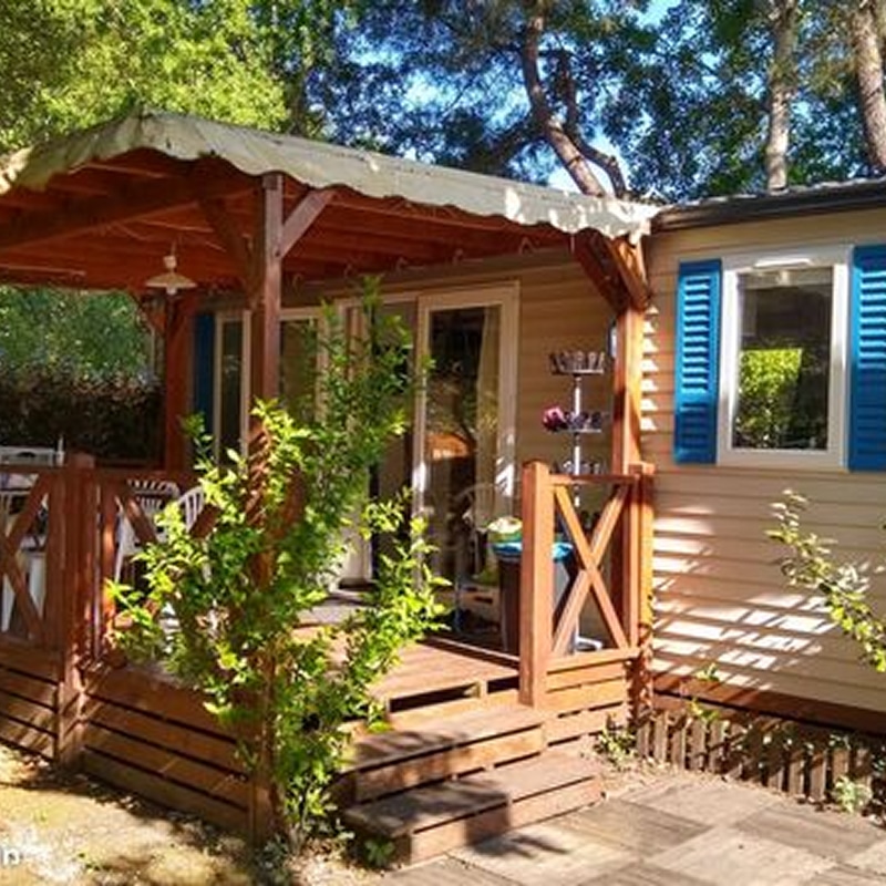 Bungalow de camping : terrasse bois et volets bleus Mobile home de camping beige dans la forêt. Grande terrasse couverte en bois, marches, portes vitrées et volets bleus éclatants.