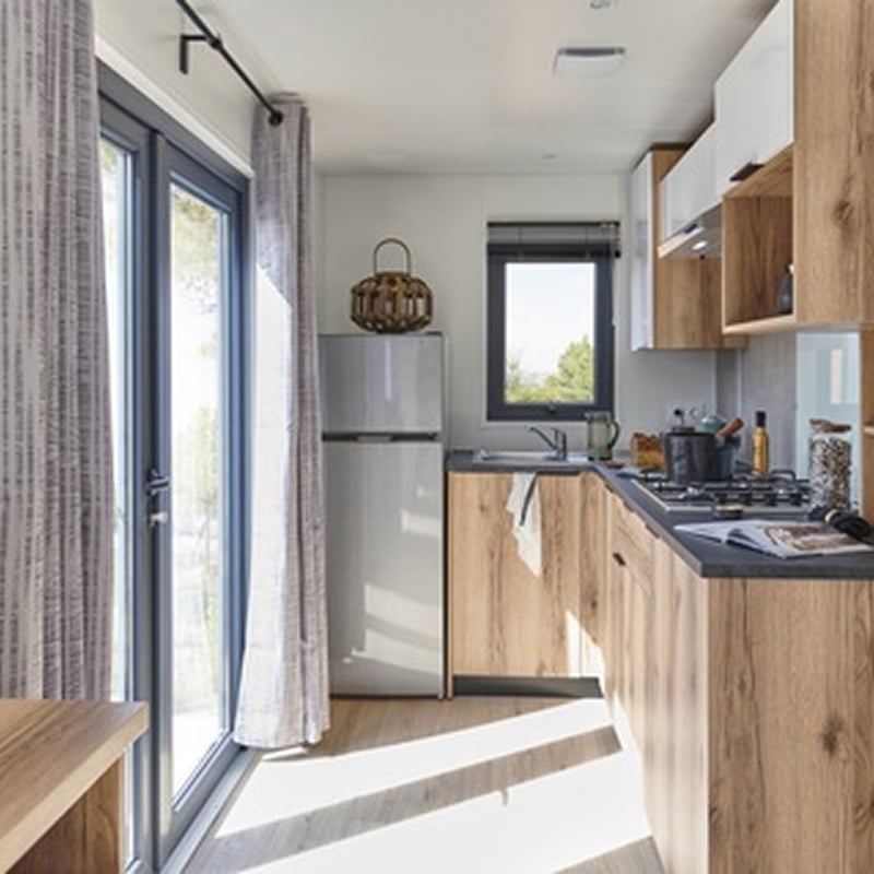 Petite cuisine compacte et lumineuse en bois clair. Cuisine moderne et compacte de mobil-home avec meubles en bois clair, plan de travail foncé, réfrigérateur et baie vitrée lumineuse.