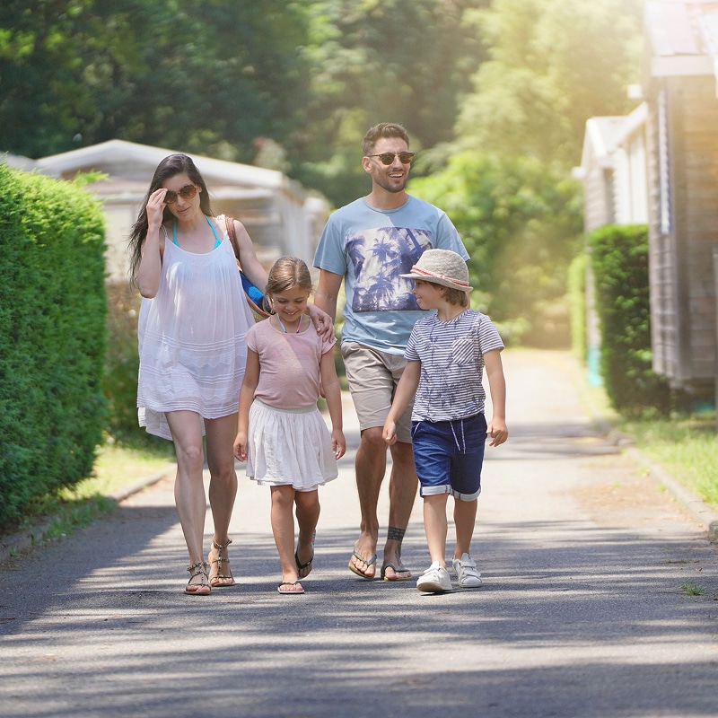 Famille en vacances d'été marchant sur un chemin ensoleillé Parents et deux enfants souriants marchent ensemble sur un chemin ensoleillé bordé de haies et de mobil-homes, profitant des vacances d'été.