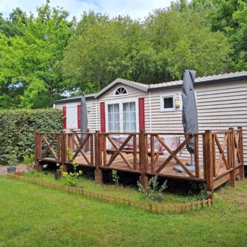 Mobil-home et terrasse en bois, cadre parfait pour les vacances. Mobil-home beige avec volets rouges et lucarne, doté d'une grande terrasse en bois rustique, sur un terrain gazonné en pleine nature.