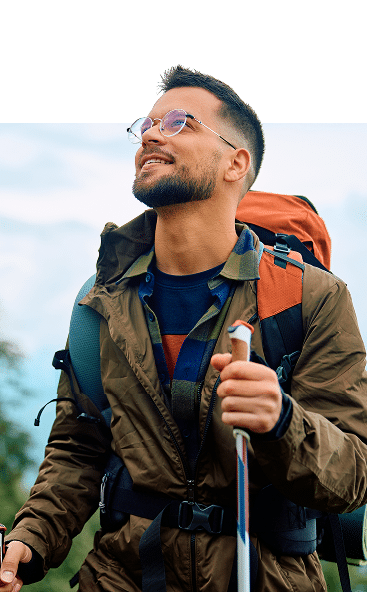 Randonneur souriant : prêt pour le trekking en montagne Randonneur souriant avec lunettes, veste marron et chemise à carreaux. Il porte un sac à dos orange et tient des bâtons de trekking.