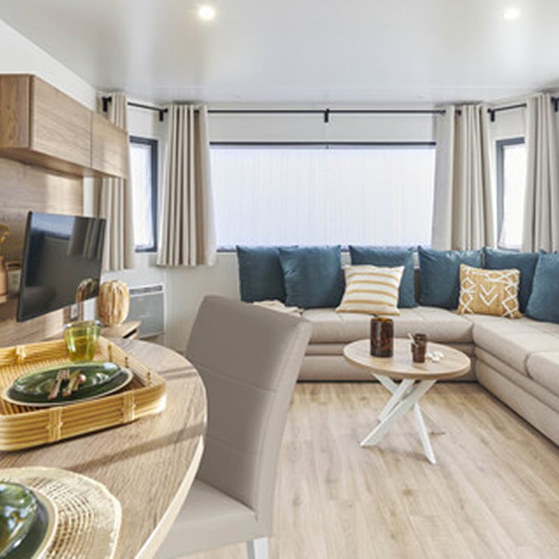 Canapé d'angle et salon design pour un lodge moderne Salon moderne d'un lodge: canapé d'angle beige, coussins bleu sarcelle, table basse ronde et coin TV/repas. Intérieur lumineux et design.