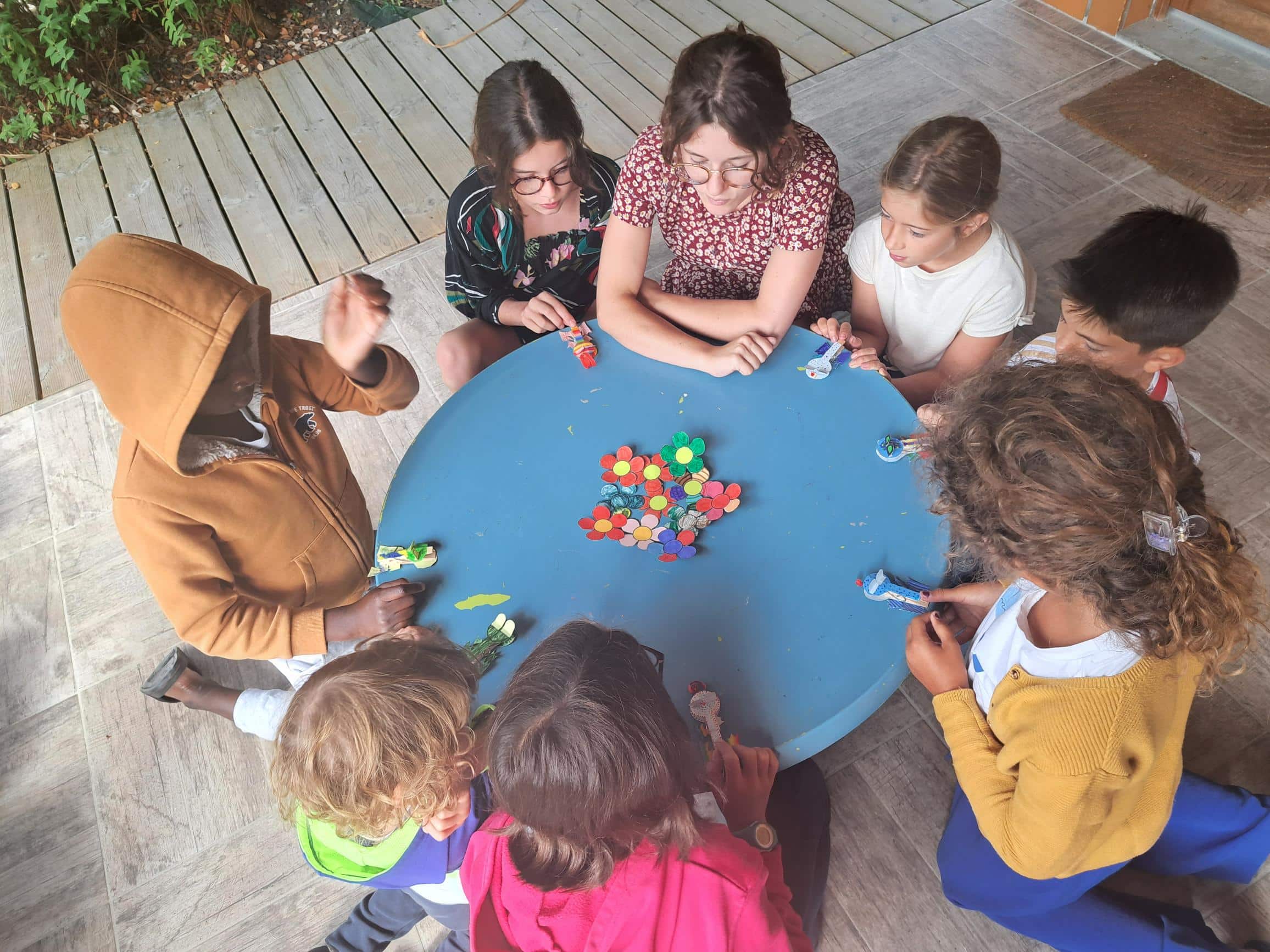 Enfants et animateurs réunis autour d'une table bleue pour un atelier créatif de fleurs en papier en extérieur.