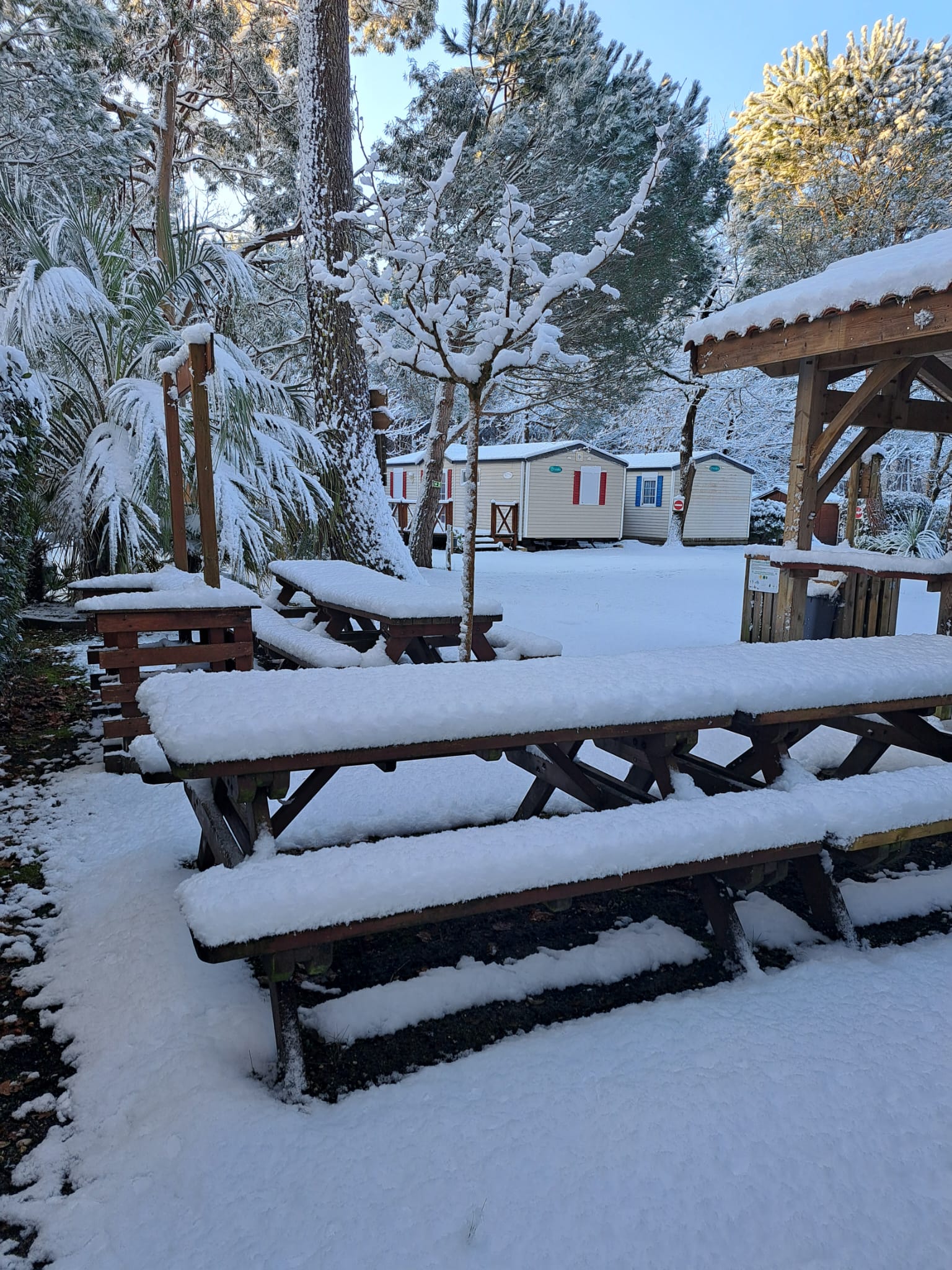 Tables de pique-nique sous la neige épaisse dans un camping avec mobil-homes et arbres enneigés par un matin d'hiver.