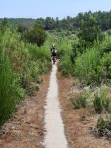 Deux cyclistes parcourent un étroit sentier blanc entouré d'une végétation dense et de pins sous un ciel clair.