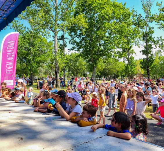 Enfants attentifs devant un spectacle en plein air à L'Eden Aquitain sous les arbres ensoleillés.