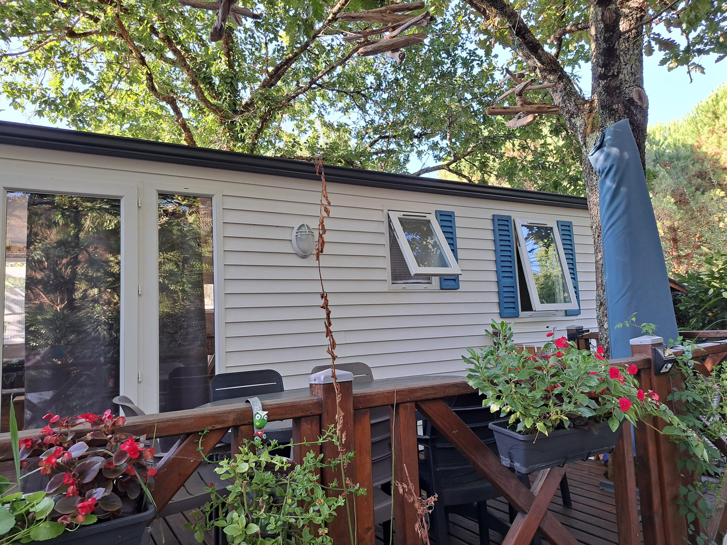 Mobil-home blanc avec terrasse en bois fleurie et volets bleus sous les arbres.
