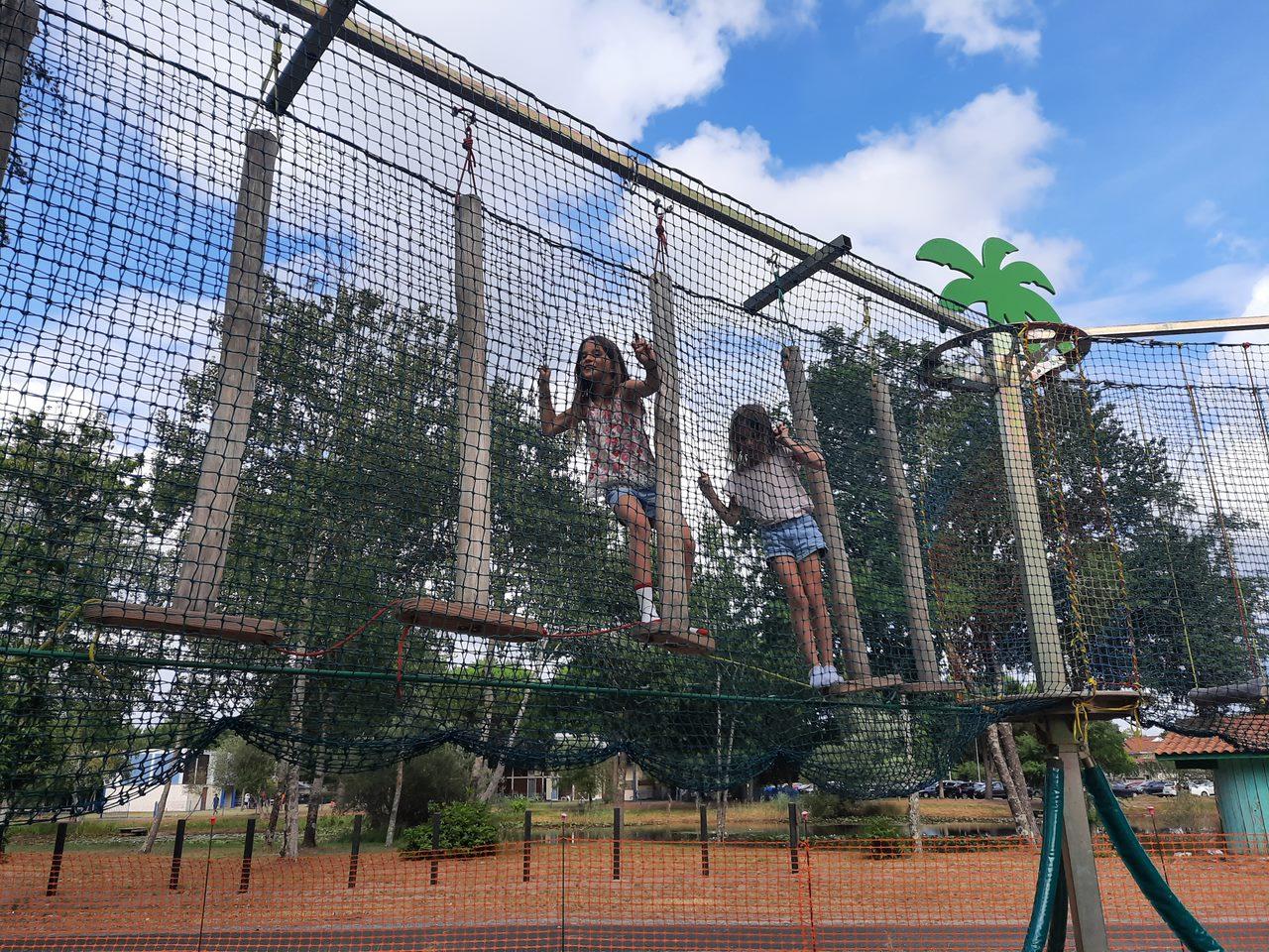 Deux fillettes s'amusent sur un parcours d'accrobranche sécurisé par des filets lors d'une journée ensoleillée.