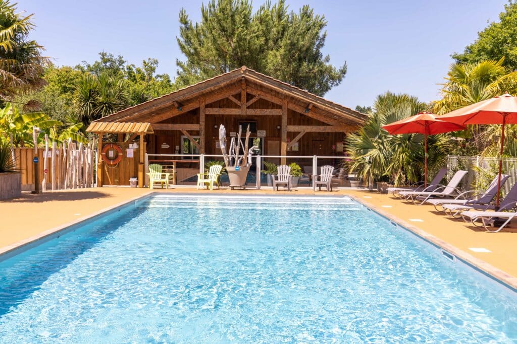 Piscine avec chalet en bois et espace détente arboré Piscine ensoleillée devant un chalet en bois avec transats, palmiers et panneau réception.