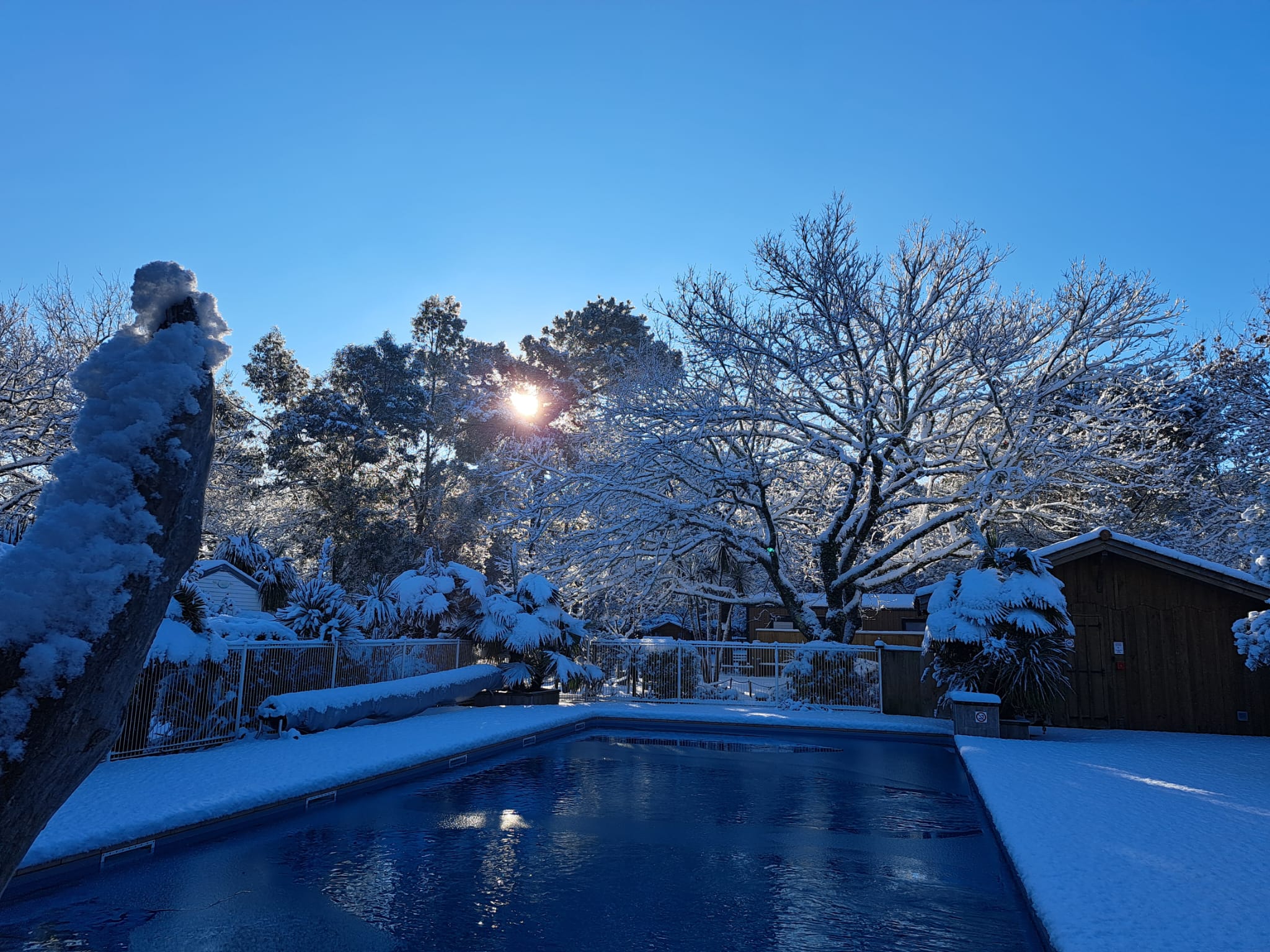 Piscine sous la neige avec des arbres givrés et un soleil éclatant sous un ciel bleu d'hiver pur.