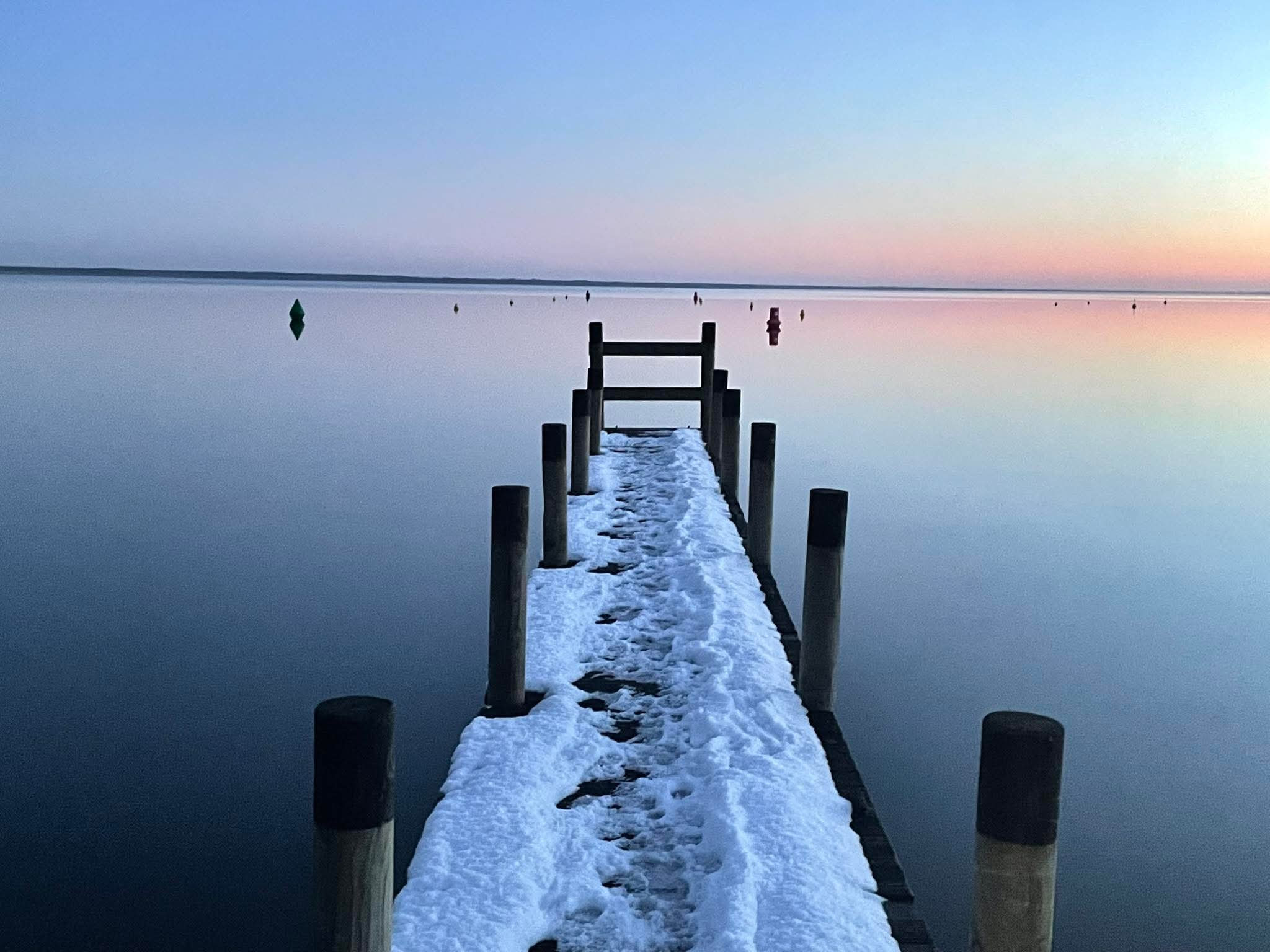 Ponton en bois enneigé s'avançant sur un lac calme au crépuscule avec des bouées sous un ciel pastel.