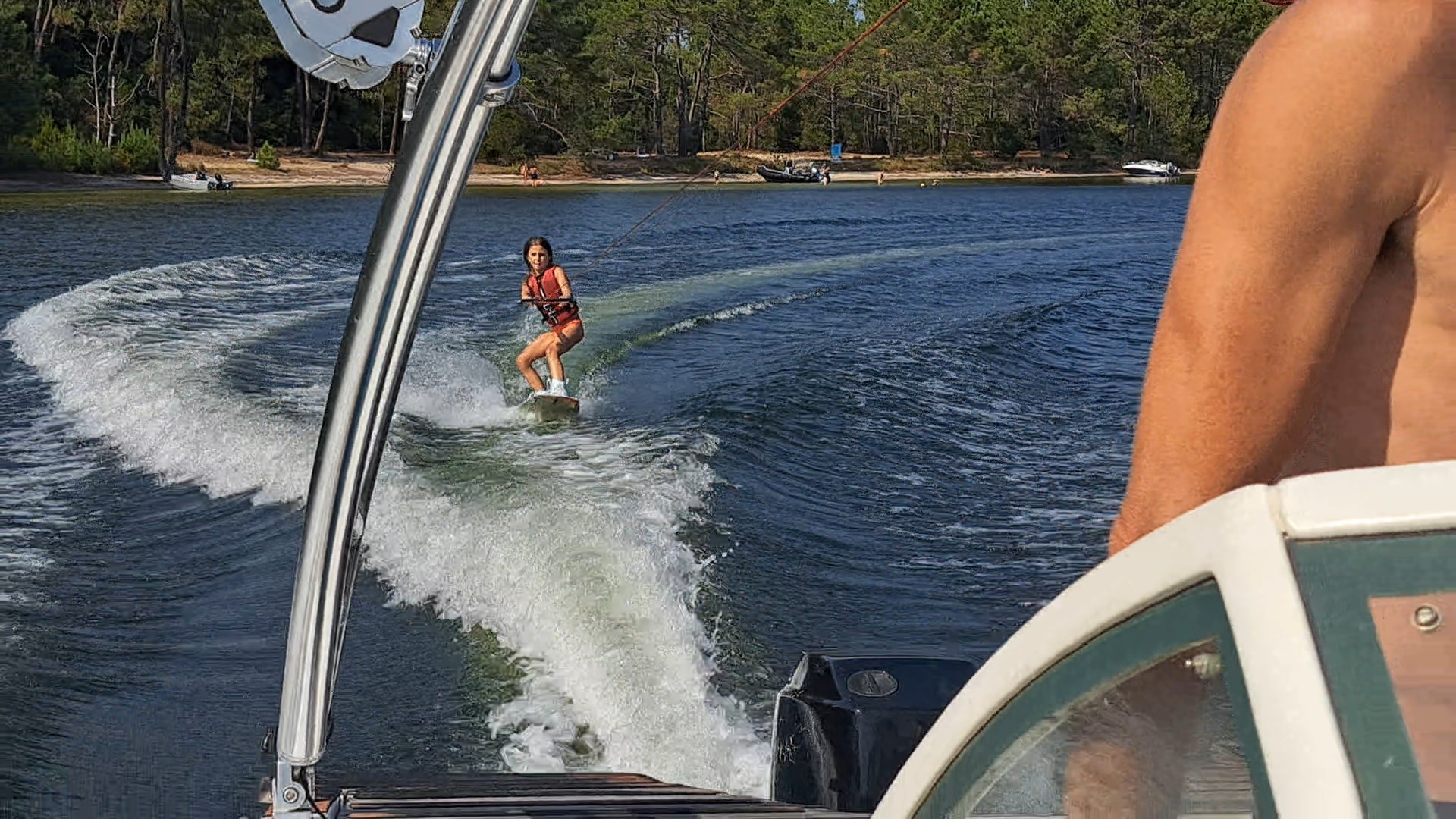 Jeune fille en gilet orange faisant du wakeboard sur un lac paisible bordé de pins au soleil.