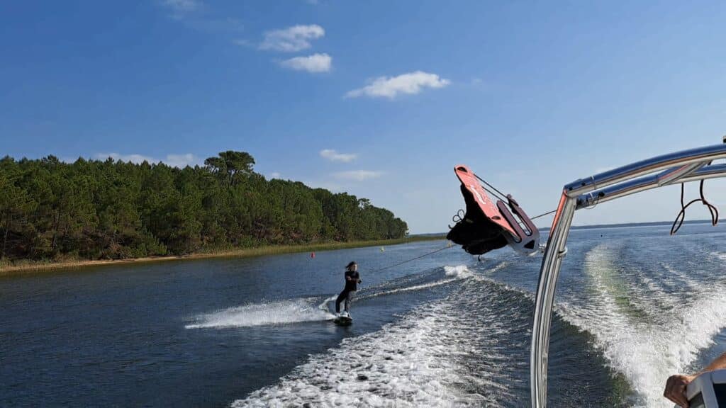 Une personne pratique le wakeboard sur un lac paisible bordé d'une forêt de pins sous un ciel bleu.